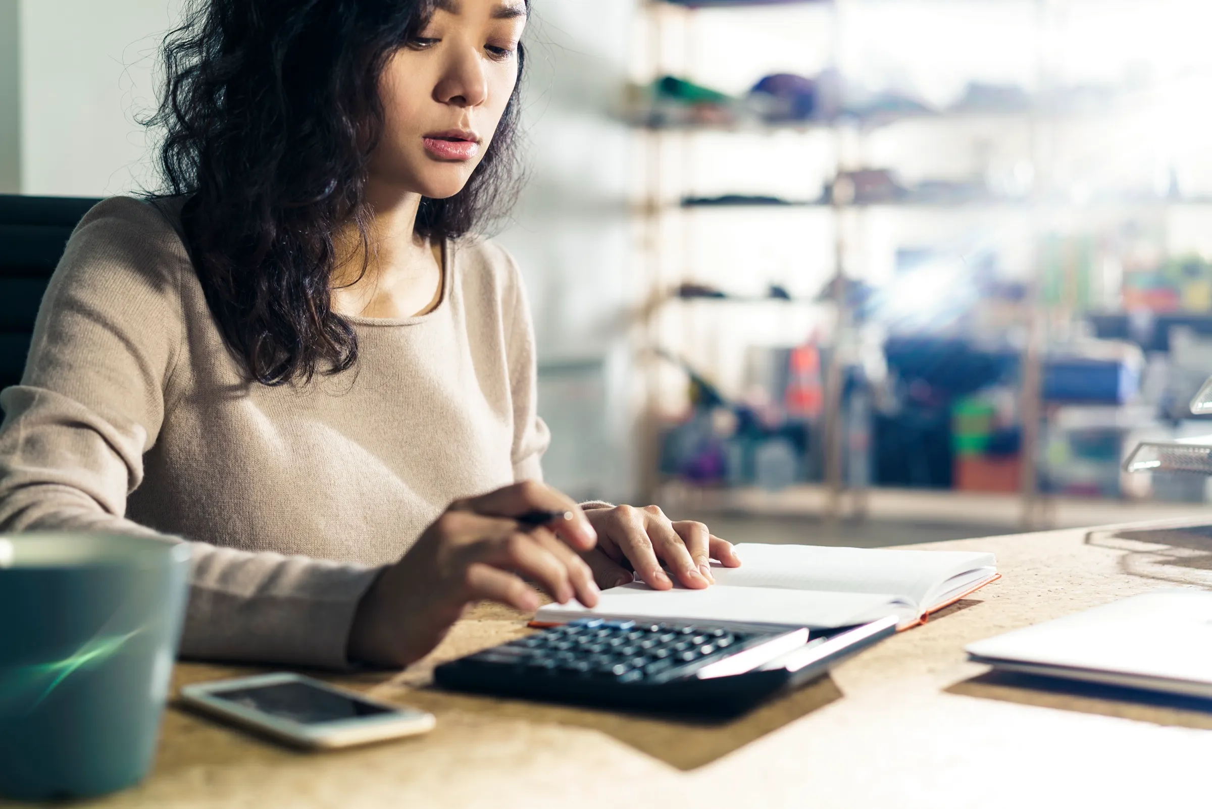 Antreprenoare calculând bugetul lunar de publicitate, cu calculator, agendă și laptop pe birou